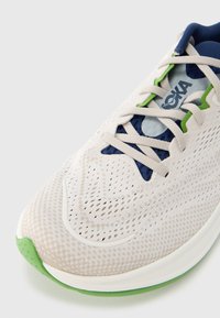 Chaussure de course légère avec une tige en mesh blanc cassé, des accents bleu marine et une semelle verte. Présente un tissu texturé et des lacets plats.