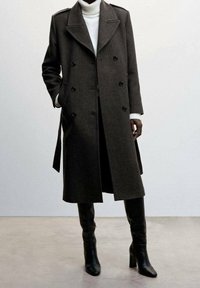 Manteau en laine brun à double boutonnage avec de gros boutons, épaulettes et poches latérales, associé à un col roulé blanc et des bottes hautes noires jusqu'au genou.