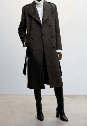 Manteau en laine brun à double boutonnage avec de gros boutons, épaulettes et poches latérales, associé à un col roulé blanc et des bottes hautes noires jusqu'au genou.