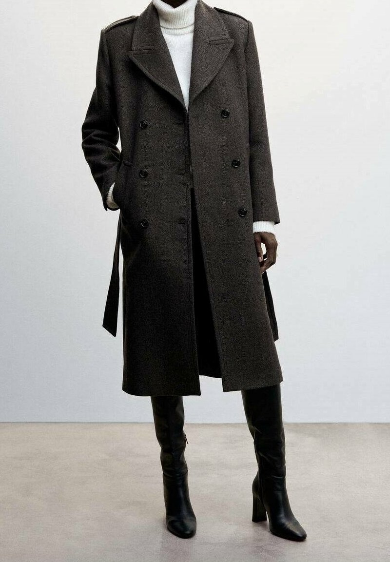 Manteau en laine brun à double boutonnage avec de gros boutons, épaulettes et poches latérales, associé à un col roulé blanc et des bottes hautes noires jusqu'au genou.