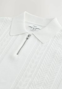 Polo de punto blanco con cierre de cremallera, que presenta patrones de diamante texturizados a los lados y un cuello acanalado. Etiqueta de marca visible.
