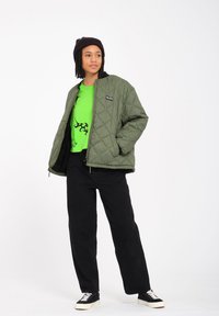 Oliven grøn quiltet jakke med lynlås, parret med en lys grøn grafisk t-shirt og sorte brede bukser. Sorte sneakers med hvide detaljer.