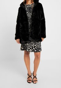 Černý kabát z umělé kožešiny s kapucí, s hladkou texturou a předním zapínáním. Nosený přes vzorované černobílé šaty.