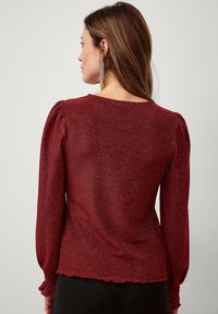 Bordeaux kleurige blouse met lange mouwen, een textuur met glinsterende afwerking, gepofte schouders en smock banden. Ronde zoom met een gerimpelde rand.