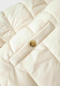 Detalle de una chaqueta tipo puffer crema con un botón dorado y un texturizado acolchado. La tela es suave y ligeramente brillante, con un brillo sutil.