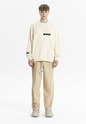 Hombre de pie con sudadera beige oversize con texto "MWM", pantalones beige a juego y zapatillas beige texturizadas sobre fondo blanco.