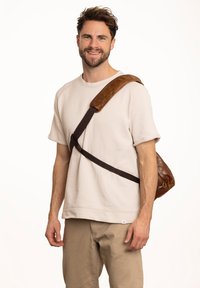 Brown leather messenger bag with a wide padded strap, textured finish, and a keyring attachment, worn across the body against a light backdrop.