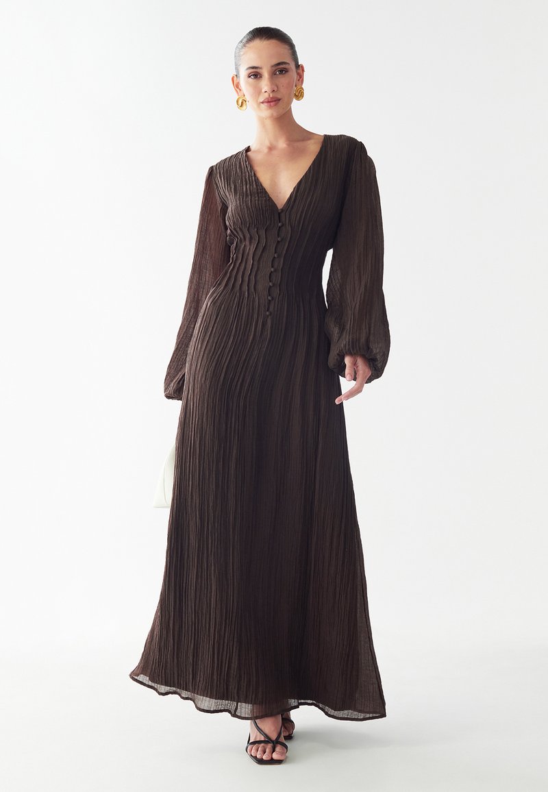 Dolga, rjava plisirana obleka z V-izrezom, sprednjim zapenjanjem na gumbe in napihnjenimi rokavi. Gladka tekstura z tekočo silhueto.