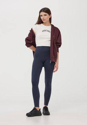Felpa con zip color bordeaux, crop top bianco con testo "NEW YORK", leggings blu scuro e sneakers nere. Tessuti morbidi e design aderente.