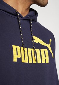 Felpa con cappuccio blu navy con un audace logo "PUMA" giallo e un motivo grafico. Presenta spesse corde di regolazione con dettagli gialli. Tessuto morbido.