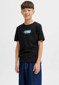 Nasmejani dječak u crnom Jack & Jones majici i širokim plavim trapericama, stoji pored jednostavne svijetle pozadine.