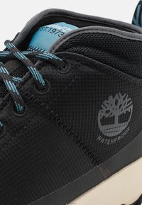 Zapato negro impermeable con un upper texturizado, cordones azules y un logo con un diseño de árbol. Incluye un collar acolchado para mayor comodidad.