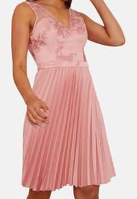 Rosa plisserad klänning med blommönsterbroderi, v-ringning, genomskinliga detaljer och en figurnära bodice. Tyget har en mjuk lyster och en strukturerad look.