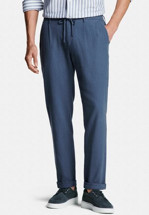 Homme portant un pantalon bleu à cordon, revers roulés, baskets bleu marine à semelles blanches et une chemise rayée à manches longues, debout les mains dans les poches.