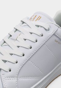 Baskets blanches GAP avec lacets blancs, logo "GAP" doré sur la languette et détails cousus sur la tige en cuir lisse.