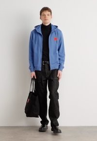Blå hettejakke med glidelås og rødt logo, svart turtleneck, mørke bukser, svarte sko og en svart tote-bag. Glatt tekstur, avslappet passform.