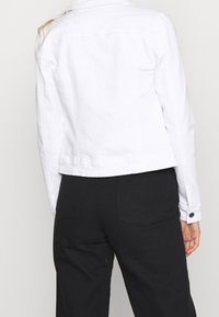 Veste en denim blanc à coupe courte, avec poignets à boutons et découpe sur le dos, associée à un pantalon noir doté de poches plaquées.