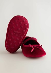 Pehmeät punaiset vauvan kengät, joissa on pieni nauha, yksi kengistä näyttää pohjan, jossa on kohokuvioituja sydämiä ja teksti "NEXT baby".