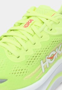 Φωτεινό κίτρινο-πράσινο παπούτσι τρεξίματος HOKA με κορδόνια, άνω μέρος από διαπνέον πλέγμα και λευκή σόλα με το λογότυπο της μάρκας στο πλάι και στη γλώσσα.