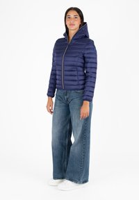 Marineblauwe pufferjack met capuchon, met een ritssluiting aan de voorkant, een horizontale quiltstructuur en elastische manchetten. Gemaakt in combinatie met wijde jeans en witte sneakers.