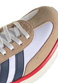 Primer plano de una zapatilla deportiva de ante beige y malla blanca con rayas azules marinas, cordones blancos y suela con detalles en rojo.