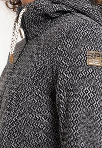 Giacca grigia e nera a motivo con chiusura a zip, cappuccio regolabile, coulisse e una piccola toppa con il marchio sulla manica.