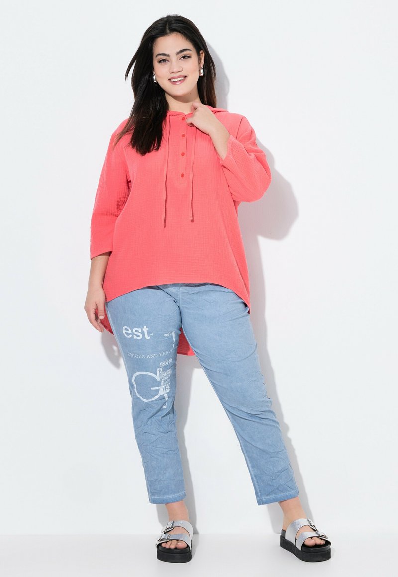 Blusa corallo oversize con maniche a tre quarti, bottoni frontali e cappuccio con cordoncino. Pantaloni azzurri chiari con grafiche stampate e vestibilità rilassata.