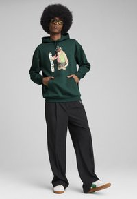 Tamsiai žalia džemperis su priekine kišene ir grafika, vaizduojančia figūrą, laikantį snieglenčių. Juodos plačios kelnės ir baltos sportbačiai.