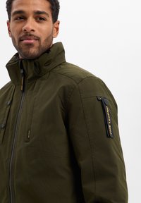 Olijfgroene jas met een hoge kraag, rits en knoopsluiting, meerdere zakken en een logopatch op de mouw. Gemaakt van gladde stof.