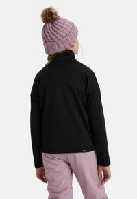 Černá fleecová bunda s vysokým límcem a texturovanou tkaninou, nošená s světle růžovými kalhotami. Pletená světle fialová čepice s kožešinovou bambulí.