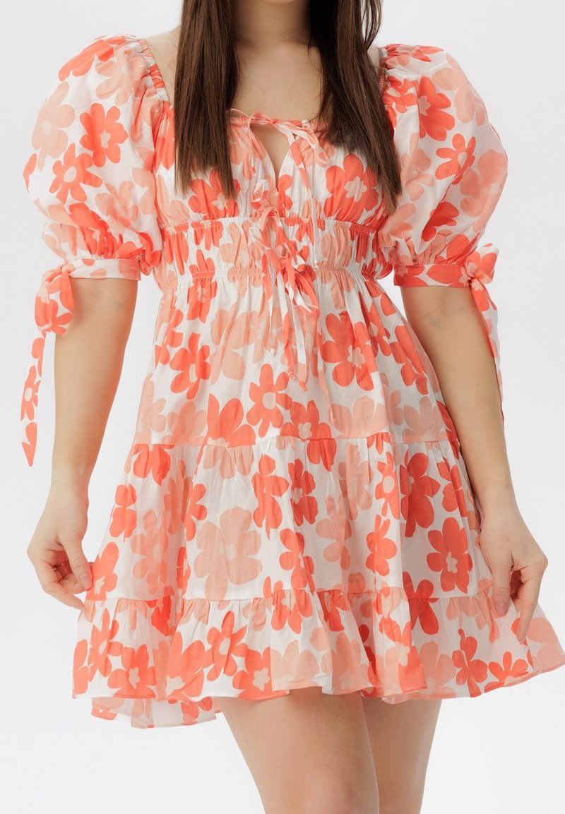 Femme portant une robe courte blanche avec un imprimé floral orange, des manches bouffantes nouées aux coudes et un décolleté en trou de serrure avec des liens à cordon.
