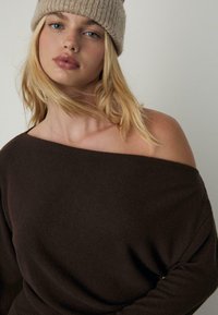 Braunes, schulterfreies Sweatshirt aus weichem Material in lockerer Passform. Modell trägt eine hellbeige, gerippte Strickmütze.