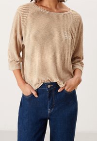 Gestreiftes, beiges und braunes Langarmoberteil mit lockerer Passform und Rundhalsausschnitt, kombiniert mit dunkelblauen Jeans und Händen in den Taschen.