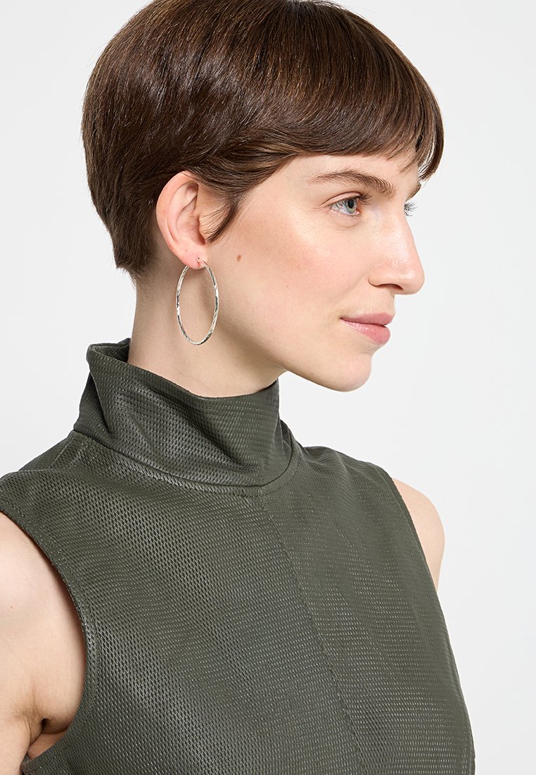 Top de alto cuello en un elegante tono verde oliva con un patrón texturizado, combinado con pendientes grandes de aro plateado. Cabello corto peinado de forma ajustada.