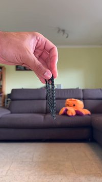 Melna metāla ķēde turēta rokā, ar savienotām acīm. Fons ietver brūnu dīvānu un oranžu pūkainu astoņkāju.