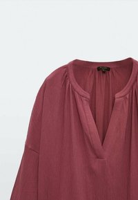 Blouse ample à manches longues de couleur rose poussiéreuse avec tissu froncé au niveau de l'encolure et col ouvert en forme de V.