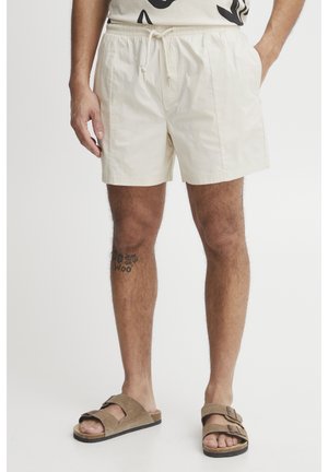 Beige short swim trunks with elastic waistband and drawstring. Lightweight fabric, side pockets, paired with brown slide sandals.