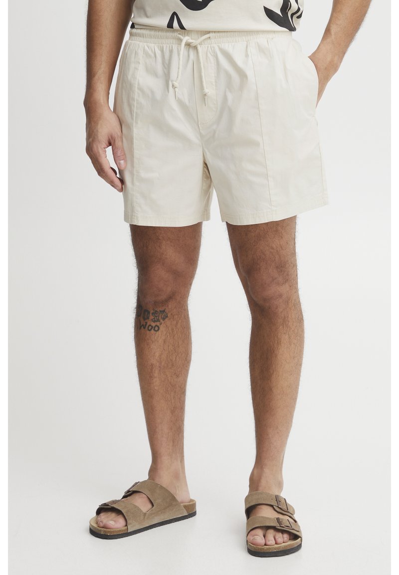 Beige short swim trunks with elastic waistband and drawstring. Lightweight fabric, side pockets, paired with brown slide sandals.