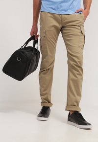 Bolso de lona negro con dobles cremalleras, pantalones cargo color caqui con bolsillos laterales, y zapatillas negras con suelas blancas. Camiseta de color azul claro visible.