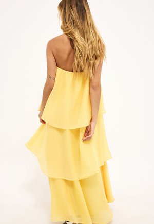 Femme aux longs cheveux blonds portant une robe jaune à volants sans bretelles, debout de dos contre un fond uni.