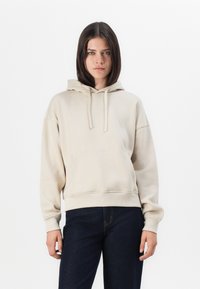 Sweat à capuche beige en tissu doux, avec une poche kangourou, un capuchon à cordon et des poignets et un ourlet côtelés. Associé à un jean foncé.