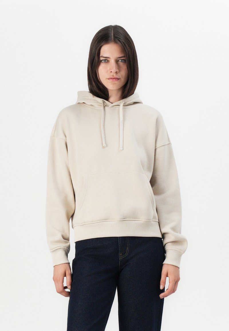 Sweat à capuche beige en tissu doux, avec une poche kangourou, un capuchon à cordon et des poignets et un ourlet côtelés. Associé à un jean foncé.