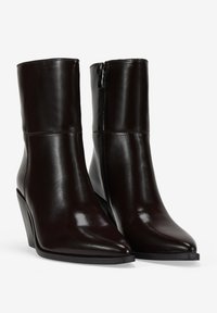 Botas de cuero marrón hasta el tobillo con una textura suave, punta afilada y tacón curvado; cuentan con un cierre de cremallera en el lateral.