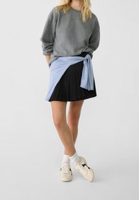 Sweatshirt gris avec épaules dénudées, enroulement bleu autour d'une jupe plissée noire, et baskets crème avec accents noirs, chaussettes blanches visibles.