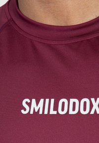 Burgundy idrottströja med rund halsringning, tillverkad av slätt, åtsittande tyg med den vita texten "SMILODOX" framträdande.
