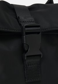 Zwarte roll-top tas met een stevige plastic gesp, gestructureerde riem en gladde stof, met een minimalistisch ontwerp en functionele accenten.