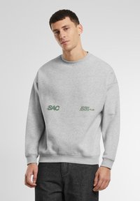Hanorac gri cu guler rotund, fabricat dintr-un amestec de bumbac, având text verde „SAC” și „SATURDAY ATHLETICS CLUB” pe partea din față. Manșete și tiv din material cusut.