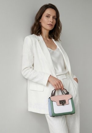 Vrouw in een volledig wit pak staat tegen een grijze muur, en houdt een kleine rechthoekige handtas met roze, witte, groene en blauwe panelen vast.