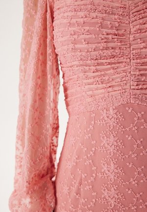 Robe rose avec un corsage en tissu froncé, broderie florale texturée, manches longues transparentes et une silhouette fluide.