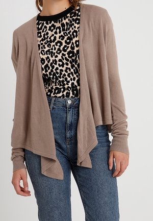 Femme portant un cardigan beige ouvert devant sur une chemise à imprimé léopard noire et beige, associée à un jean bleu taille haute.